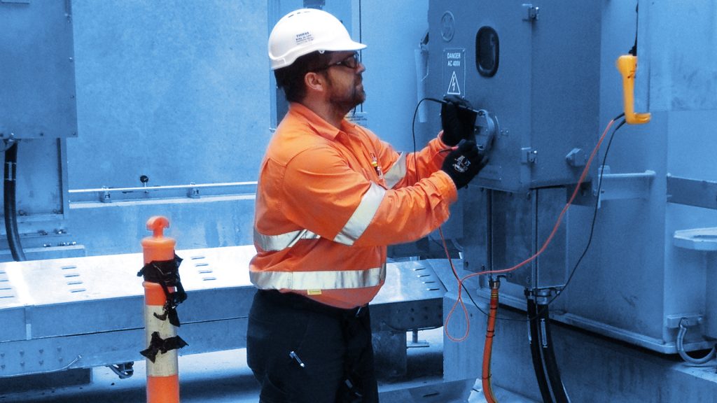 Profile of Westlake Environmental's Field Service team member, doing a control panel test, orange safety jacket and safety cone, background blue wash